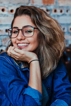 A young woman wearing glasses and denim, smiling in an indoor setting.